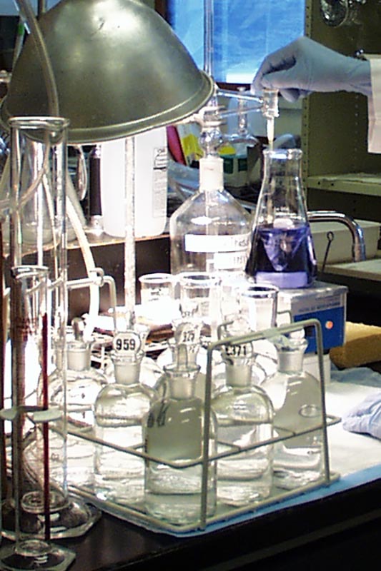 A rack of glass bottles with stoppers sits on a counter near a flask-mixing unit. A blue-gloved hand adds a purple substance to the mixture in the flask on the unit.