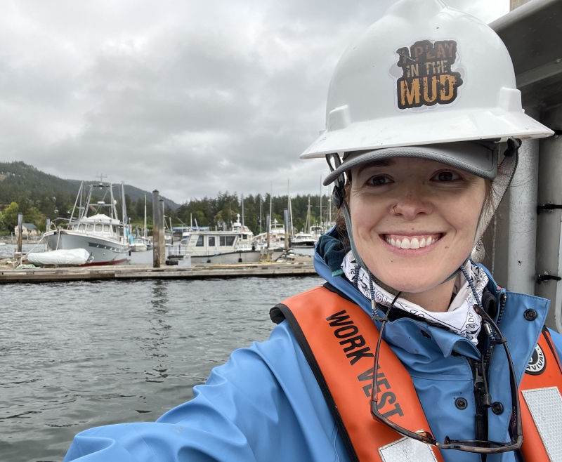 Selfie of a smiling scientist in a hard hat and life jacket, on the back deck of a boat on a cloudy day. Other boats are visible in the background.