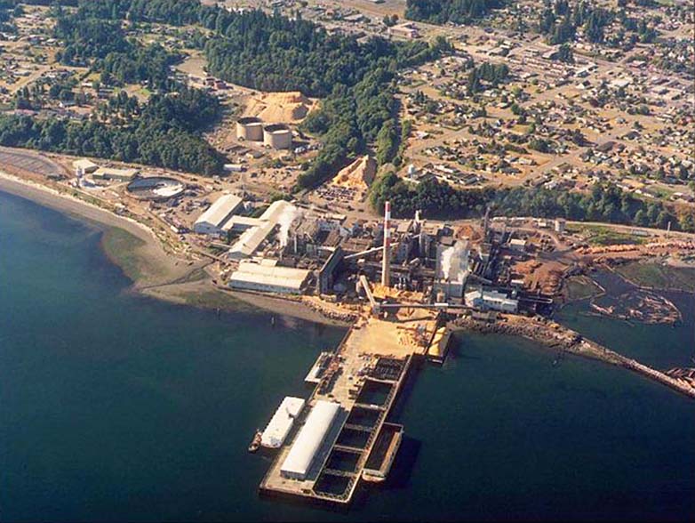 Aerial view of shoreline with large industrial buildings and smokestacks. A very large wooden pier juts into the water and logs float in a lagoon behind a jetty.