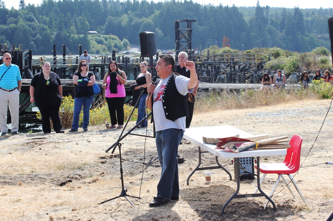 Man speaking with microphone in front of crowd, holding left hand in air with a closed fist.