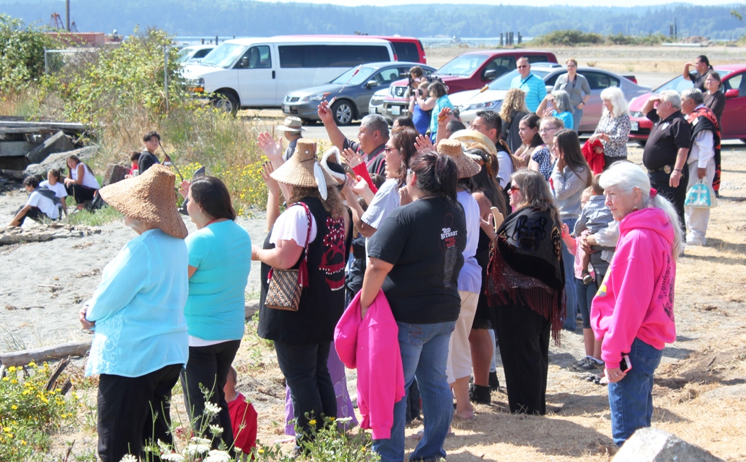 Crowd standing and watching canoe event from the sandy shore.