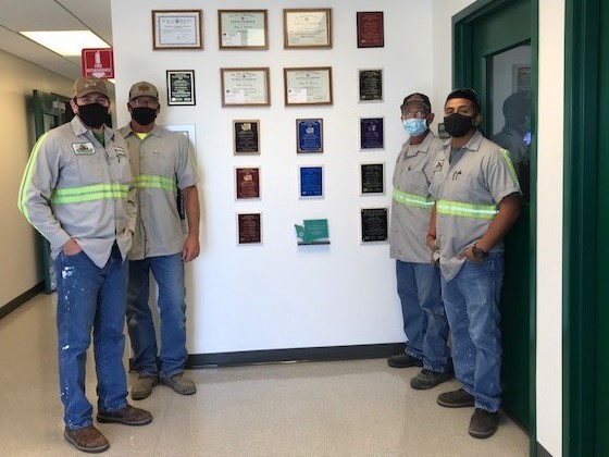 Wastewater Treatment Plant operators stank proudly next to a wall covered in awards wearing covid-19 masks