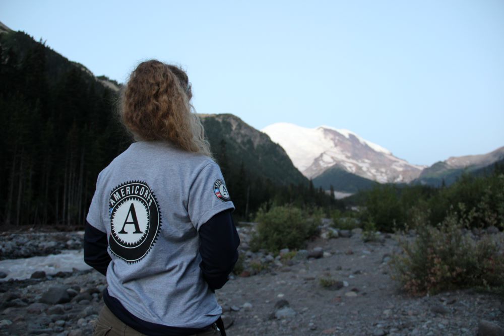 A WCC member stands with their back to the camera, facing a mountain in the distance. Their shirt has a large AmeriCorps logo on the back.
