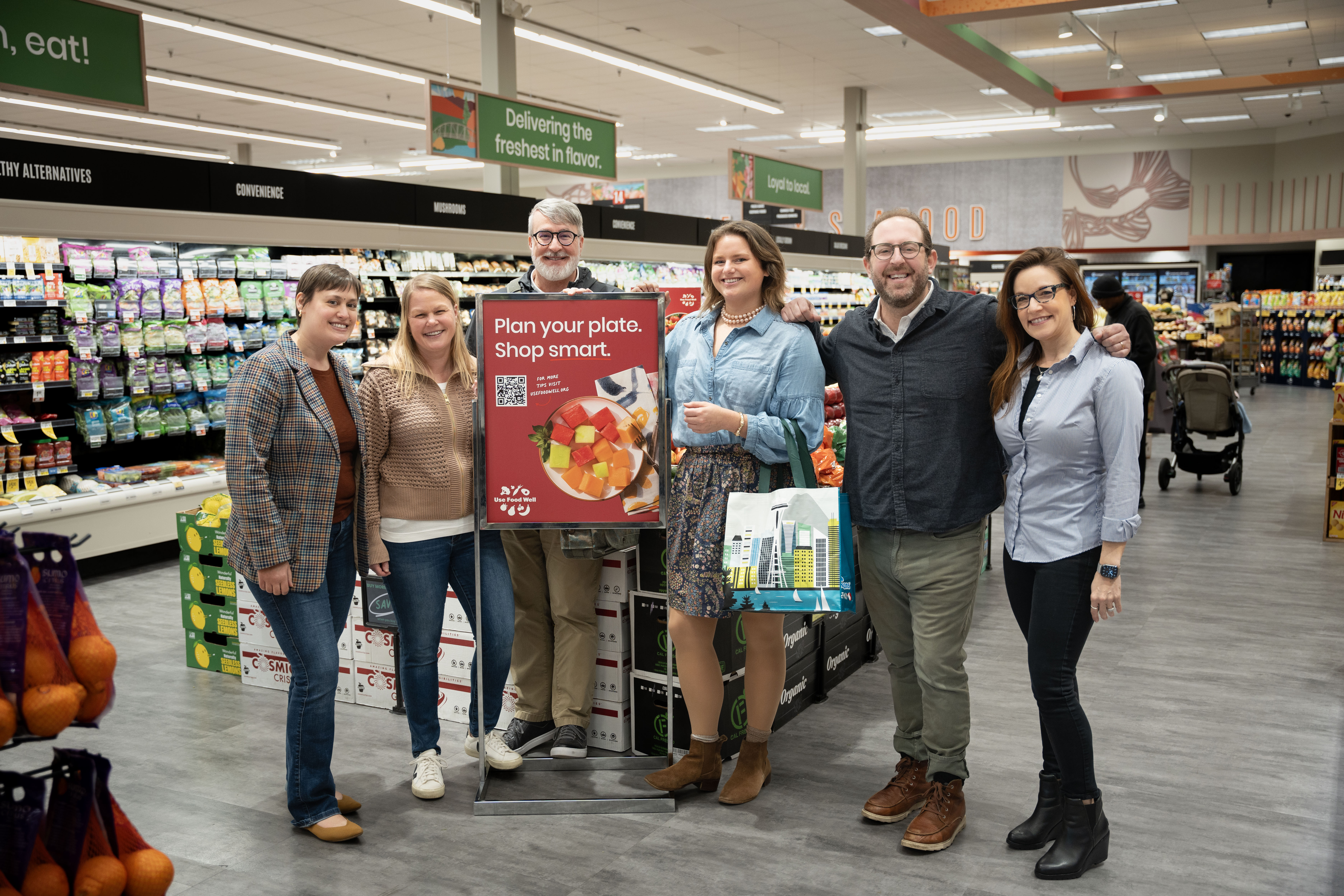 Six smiling adults in grocery store around sign that says 